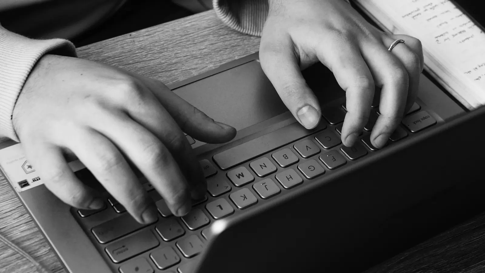 يدان تكتبان على لوحة مفاتيح حاسوب محمول بجانب دفتر ملاحظات مكتوبة بخط اليد